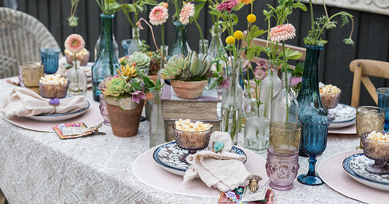 An image of a purple, blue and light pink glasses and dishes dining set up surrounded by dahlias and succulents