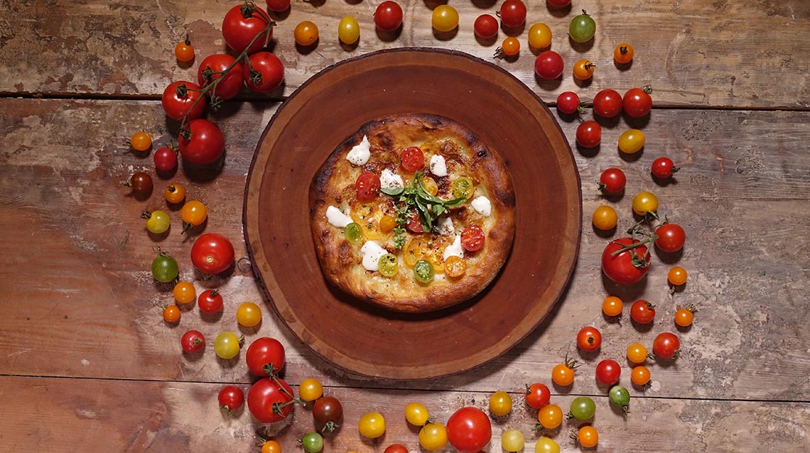 An image of a personalized mozzarella and tomato pizza with a variety of garden tomatoes surrounding it placed on a wood table.