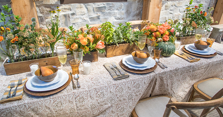 An image of a fall table that is set with a cream color tablecloth, rattan placemats, flowers in shades of oranges and herbs and pears.